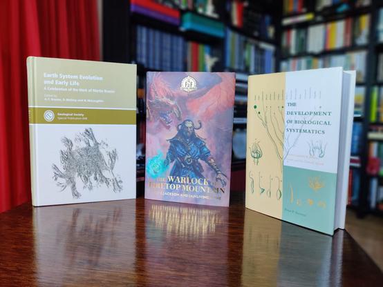 A photo of three hardback books standing upright on a small, brown, wooden table. The out-of-focus background shows black shelves full of books and part of a red curtain covering a door

On the left, Earth System Evolution and Early Life, showing a black and white line drawing of a fossil at the bottom half, an ochre-coloured band across the middle with the publisher's name, and a cream-coloured top section with the title. It's hard to make out what the fossil is; it appears as a black square with several bits sticking out on all sides, but the legend explains it is a camera lucida drawing of a fossil stauromedusan, Haootia quadriformis (a species of stalked jellyfish), from the Ediacaran of Newfoundland, Canada.

In the middle, The Warlock of Firetop Mountain, showing a colour painting of an old male sorcerer in a blue coat with a flaring collar. His right hand is enveloped in a bright-blue plasma. A large, winged, red dragon looms in the background. The title is stamped across the bottom in gilded letters.

On the right, The Development of Biological Systematics, showing several line drawings in two-tone colours (cream and green) of plant parts and a tree-like diagram drawn in an old-fashioned style to appear like branching coral. The cover is divided into a cream left half, a white section top right, and a lime-green section bottom right.