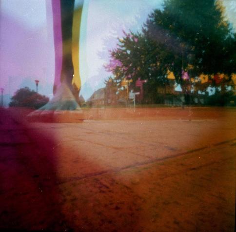 A statue of a giant bare foot and leg, shot from a low point of view.  Pavement in front, a big tree in the background. It's three layers of the same image, slightly offset, one pinkish, one yellow, and one regular colour.