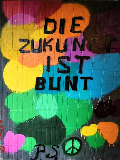 Viele farbige Flächen auf einer Wand. Darauf steht in schwarzer Farbe: DIE ZUKUNFT IST BUNT P.S. <Peace-Symbol>