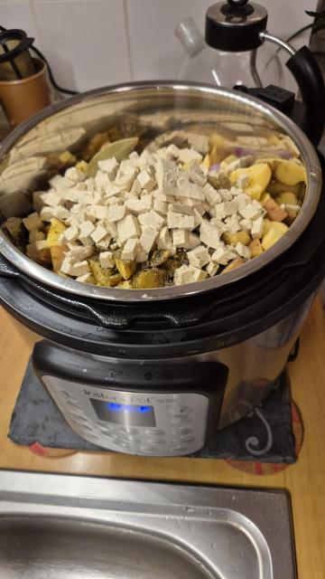 Ein automatischer Dampfkocher von schräg oben, der Edelstahleinsatz ist fast randvoll mit feschnippeltem Gemüse gefüllt, bedeckt von gewürfeltem Tofu, im Hintergrund ein Wasserkocher ubd eine weiß gekachelte Wand