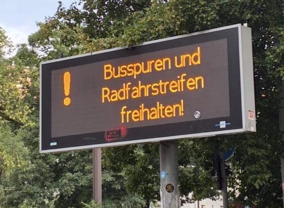 Foto von elektronischer Anzeige neben Hauptstraße. Sie zeigt den Text „Busspuren und Radfahrstreifen freihalten“