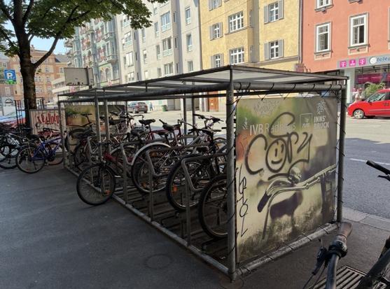 Foto von überdecktem Radparkplatz in der Größe eines Autoparkplatzes. Es stehen um die 10 Räder da. Er ist aus einfachen Rohren gebaut, mit Planendach.