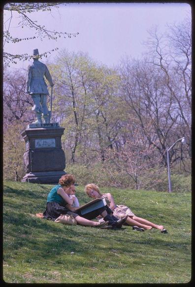 Two women sit on a grassy hill near an older statue, one reading from what looks like a box or briefcase and the other lying back with her hands under her head. There is another woman sitting behind them wearing a green top; she holds something in her arms but it's unclear if it's food or perhaps clothing. In the distance are some trees that have started to blossom, suggesting springtime activity, possibly Easter as suggested by their clothing and posture. The statue seems of an older man with his right arm raised holding what appears like a hat stand over one shoulder while walking on another foot in boots which is reminiscent of military or colonial dress.