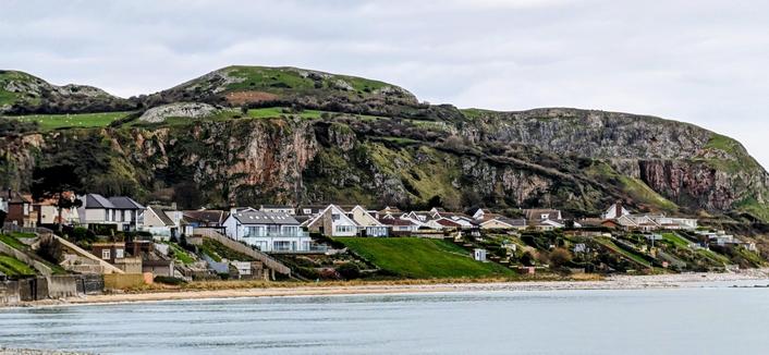 A wide shot of Penrhyn Bay, North Wales. Modern and traditional houses line the steep grassy slope facing the sea, with a rugged, green limestone headland (the Little Orme) rising dramatically behind them under an overcast sky. A narrow strip of beach meets the calm water in the foreground.

[enhanced with editing in Google Photos]