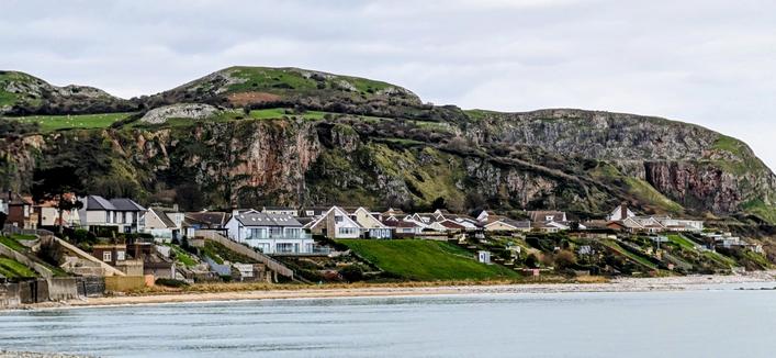 A wide shot of Penrhyn Bay, North Wales. Modern and traditional houses line the steep grassy slope facing the sea, with a rugged, green limestone headland (the Little Orme) rising dramatically behind them under an overcast sky. A narrow strip of beach meets the calm water in the foreground.

[enhanced with editing in Google Photos]
