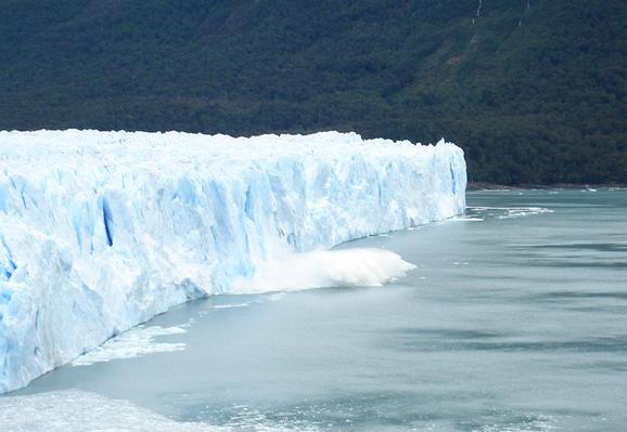 Imensa parede de gelo, no momento em que um bloco se desprendeu e caiu no lago gerando um grande espirro d'água. Ao fundo um morro com árvores.