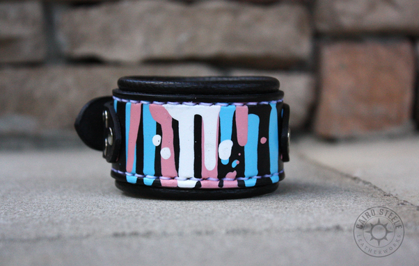 A black leather cuff with trans pride colored paint drips
