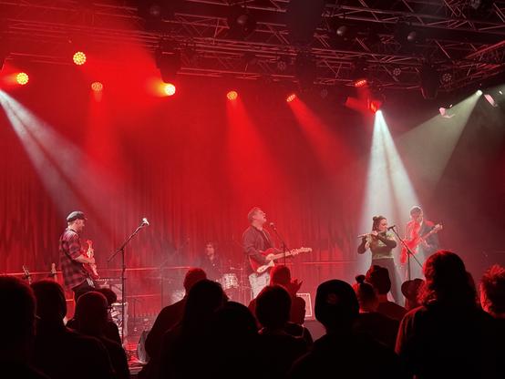 A live music performance featuring a band on stage, illuminated by red lighting. Musicians are playing various instruments, including a guitar and flute, while an audience is engaged in the foreground.