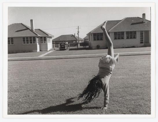 The black and white photograph captures a young child performing a handstand on a grassy lawn. The scene is set in an open, suburban area with two-story houses visible in the background under clear skies. One house has prominent windows adorned with curtains, while another features a chimney indicating possible residential use.

In the foreground, there's no discernible text or branding to suggest commercial intent. However, it appears as though this image was taken during a time when film photography was prevalent due to its grainy texture and high contrast between light and shadow areas. The absence of modern elements like digital devices suggests an earlier period in history.

The child is dressed casually, wearing what looks like comfortable trousers and a striped top with loose hair cascading down from the head's upward position during the handstand. This image conveys a moment of childhood playfulness amidst the domestic backdrop of suburban life.