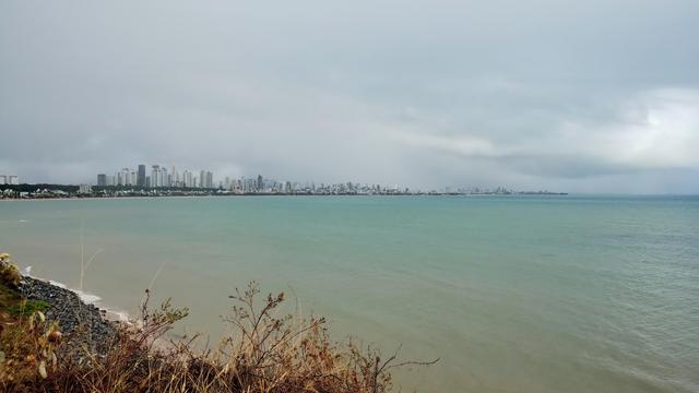 Foto tirada ao ar livre em dia de céu bem nublado. Mar de água em tom claro de azul-esverdeado. Ao fundo os prédios da região mais urbanizada do litoral de João Pessoa.