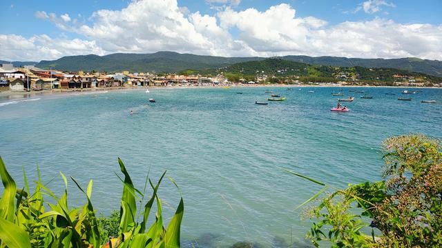 Foto tirada ao ar livre em dia ensolarado. Em primeiro plano vegetação verde e alta. Em segundo plano uma baía com água de cor turquesa claro e muitas embarcações de pequeno porte. Muito perto da linha da costa estão ranchos tradicionais. Perto deles, mais atrás, estão as casas e prédios do centro da cidade. Uma linha de muitos morros se estende no horizonte e nuvens volumosas pairam sobre eles.