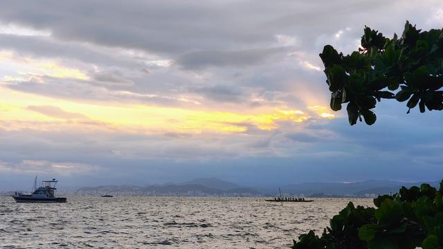 Foto tirada ao pôr do sol em tarde nublada. Em primeiro plano silhueta de galhos de árvore. Em segundo plano o mar com uma embarcação de pequeno porte mais próxima e uma canoa com alguns canoístas mais distante. O céu tem nuvens em tons de lilás e amarelo.
