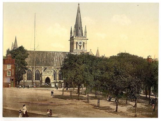 The image depicts a historic church with a prominent spire and clock tower, set against a backdrop of trees. The architecture features Gothic elements such as pointed arches and ornate tracery on the windows. In the foreground, there are people dressed in period clothing suggesting an early 20th-century setting. A few individuals can be seen walking near wooden planks or logs lying across the ground. This scene captures a moment of daily life around this significant landmark in Great Yarmouth, Norfolk, England.