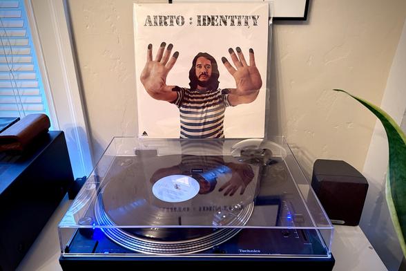 A black vinyl record spins on a black turntable. The record sleeve stands up on the turntable’s closed dust cover behind the platter