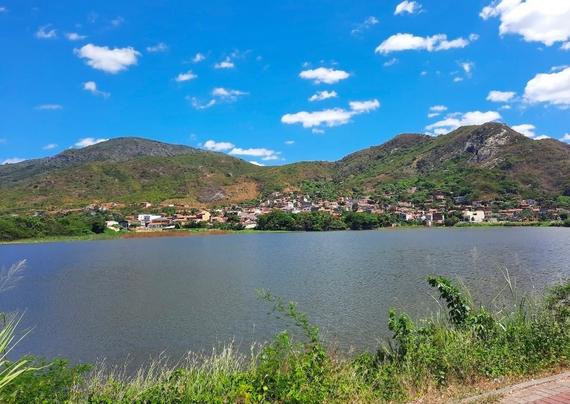 Na imagem vê-se o céu com algumas nuvens, montanhas ao fundo com casas espalhadas, e um açude.