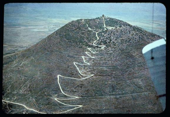The image depicts an aerial view of a winding road ascending a rocky hillside. The zigzag path suggests it might be a difficult or technical climb, possibly for vehicles navigating the terrain. At the top of the mountain is what appears to be a structure, likely religious in nature given its prominent location and apparent size relative to the surrounding landscape.

The area around the summit shows evidence of habitation or development with several buildings visible on the hillside above it. The overall setting seems arid and desolate, with sparse vegetation indicating a dry climate, possibly indicative of regions like North Africa or parts of Israel where Mount Tabor is located in biblical tradition.

In the bottom left corner, there's an overlay that appears to be part of a slide projector frame from Matson Photo Service. This suggests that the image was likely used as a visual aid for educational purposes during this time period mentioned (1950-1977), potentially related to religious studies or geography courses.

The reference "Northward from Jerusalem" might imply that the photograph is part of a series documenting various landscapes in connection with biblical narratives, specifically mentioning Mount Tabor.