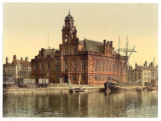 The image depicts a picturesque waterfront scene featuring an ornate brick town hall building with a prominent clock tower, located in Great Yarmouth, England. The architecture of the structure is elaborate, characterized by decorative stonework and arched windows that add to its historic charm.

Adjacent to the town hall stands a large sailing ship docked on the calm waters reflecting sunlight. Various smaller boats are also visible near the shorelines, indicating active maritime activity in this harbor setting.

The surrounding area includes several other buildings with varying architectural styles, suggesting an older urban landscape. Some of these structures feature chimneys and traditional designs typical for British seaside towns during that era.

Overall, the image captures a serene yet bustling riverside environment, showcasing the town hall as a central landmark within Great Yarmouth's historical context. The photograph is part of the Town Hall series from between ca. 1890 to ca. 1900 and reflects England’s rich maritime heritage through its depiction of urban life by the waterway.

Further details on this image can be found at [Town Hall, Great Yarmouth] (https://images.loener.nl/PhotochromPrints/full/65e4/65e47ada5890ae069f681555.jpg) via Flickr Commons project in 2015.