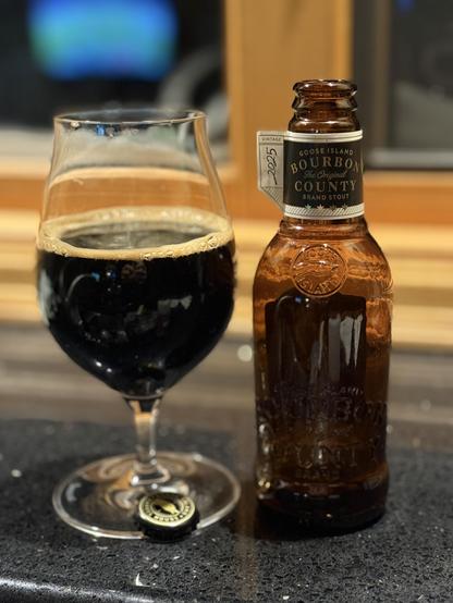 A snifter glass is half full of dark beer. The brown bottle has a black and white neck label with gold letters.  The words Goose Island and Bourbon County are embossed in the glass
