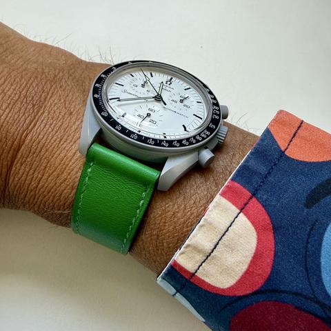 Photograph of a grey moonswatch watch with green leather band on a wrist.  The cuff of a colourful cotton shirt is visible on the right.