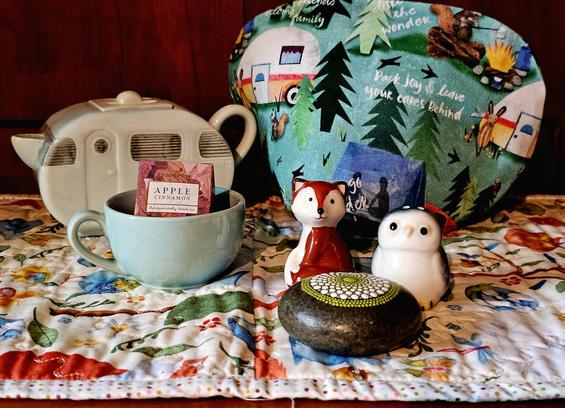 Tea tableau of a forest camp theme with an owl and a fox sitting by a firepit and in front of a teapot shaped like a vintage camper trailer. The tea is Apple Cinnamon black tea