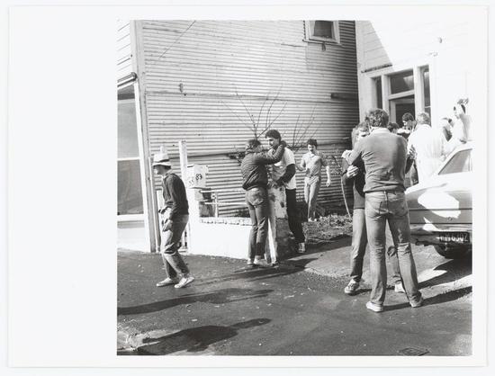 The black and white photograph captures a social gathering outside a residential building. A group of people, both men and women, is engaged in various interactions; some are embracing or dancing while others converse animatedly. The scene suggests a casual, possibly celebratory event with attendees dressed in everyday attire like jeans, jackets, and hats.
The setting includes part of the house visible on the right side, featuring windows, doors, and siding typical of suburban housing from an earlier era suggested by its style. In front is a paved surface where several individuals stand or walk around. There's also a car with a reflective white emblem parked beside one person, indicating transportation to this event.
Shadows cast across the ground suggest bright sunlight conditions during daytime hours. The overall mood seems convivial and relaxed among acquaintances or friends gathered for an outdoor social occasion in what appears to be New Zealand city life context as noted by the reference link provided at Image source.