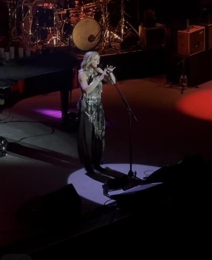 An image of Sarah on stage. She is under a soft Spotlight, singing into the microphone, her hands softly flowing near the mic. The ambient lighting behind her lights the stage in warm red and orange tones.
