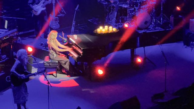 Sarah is sitting at a black grand piano, a group of lit candles atop it. The stage is bathed in blue and purple tones, and red spotlights are pointing up from behind her from under the piano.
