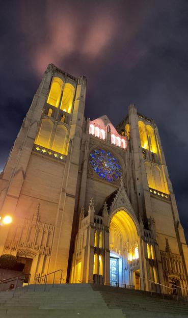 A gothic church with a tower on each side set against a foggy sky. The tower balconies are awash in a warm yellow glow. The door is awash in soft blue lights, and the round stained glass window at the center is in blue tones. The fog above the left tower is reflecting pink city lights against a dark purple grey sky.