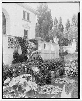 The image depicts a garden scene with classical architecture and statuary. In the foreground, there is a pond surrounded by lily pads and water plants, reflecting some of the surrounding greenery.

A stone statue resembling an Indian deity or mythological creature sits atop what appears to be a fountain structure amidst climbing vines in the background. The figure has large ears, wide eyes, and facial features characteristic of South Asian artistry. 

Behind this central garden feature is a white building with classical architectural elements such as columns and arched windows. A walled area contains more statuary and lush vegetation.

The photo appears to be black-and-white or sepia-toned, suggesting it may have been taken in the early 20th century based on its style of architecture and garden layout. The caption indicates that this image is related to the Pan American Union Building and specifically mentions a statue found within its gardens from around 1920-1950.

Overall, the scene conveys an atmosphere of cultural exchange through art, as evidenced by the presence of a South Asian-inspired figure in what seems like a Western-style garden setting.