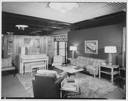 The image depicts an interior room setting with a blend of modern and traditional furniture. A piano is positioned against the left wall, accompanied by chairs arranged around it for seating during music sessions or performances. The right side features a gray couch facing away from the camera, suggesting that this area might be intended for relaxation or casual conversation rather than musical activities.

The room has several decorative elements such as paintings on the walls and patterned rugs covering large portions of the floor. Lamps provide illumination in different areas, with one near each seating arrangement highlighting both functionality and style. The overall ambiance suggests a comfortable living space designed to cater to various leisure pursuits, including music appreciation or social gatherings.

The black-and-white nature of this photograph indicates it was taken during an earlier era, reflecting design trends from the mid-20th century when such arrangements were more common in residential settings.