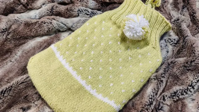 Bouillotte dans une pochette en tricot essentiellement vert tendre avec des pois blancs. Un petit pompon blanc est attaché en haut, près de l'ouverture.