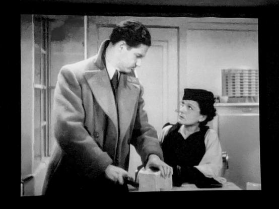 B&w scene with 2 people in a kitchen and one cutting bread