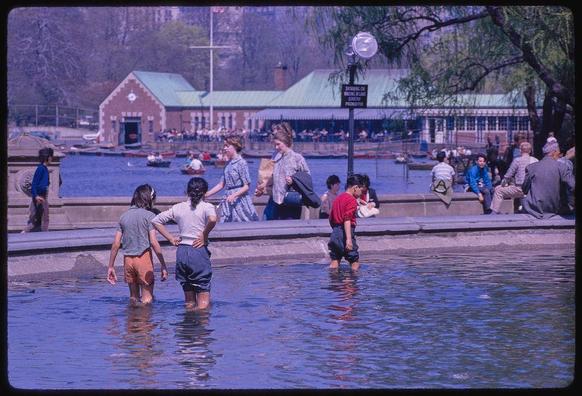 Three children wade waist deep into a public pool with one adult woman walking near them and other adults sitting along the side of the water. They are all dressed in shorts, t-shirts or sleeveless shirts and appear to be enjoying their time at Central Park on April 12th, 1962 as indicated by the caption "Central Park" under this photo description from Frissell's collection.