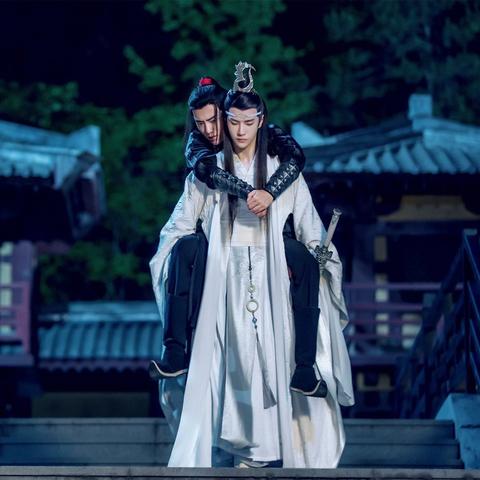 Lan Wangji passes through a city at night, bearing Wei Wuxian on his back. Wangji has Bichen in his left hand but still has both hands securely under Wuxian's thighs to help support his weight. Wuxian has his head beside Wangji's, over Wangji's right shoulder, and his arms loosely wrapped around Wangji's shoulders with his hands clasped together in front of Wangji's chest.