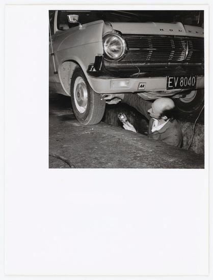 The photograph is a black and white capture of an individual engaged in mechanical work beneath the front bumper of a classic car, possibly from the mid-1960s era. The vehicle features a Holden badge, indicating it's likely Australian or British manufacture. It has round headlamps with chrome surrounds, indicative of vintage design elements popular before the widespread adoption of rectangular headlights.

The individual appears to be using an impact wrench on what could be a lug nut or wheel assembly task. They are wearing a long-sleeved shirt and trousers that suggest work attire from several decades ago. The mechanic's focus is intently directed at their tool, highlighting the manual labor aspect of auto repair during this period.

The license plate reads "EV 8040," which provides a specific detail for context but doesn't give away any personal information or exact location since it could be representative rather than unique to an individual. The setting seems informal and practical with no clear indication of professional garage facilities, suggesting the work might have been done at home or in a less formal workshop.

The image is notable as part of Hutt Valley - Mechanics collection by Ans Westra, which indicates that this photograph may belong to a larger series documenting mechanics across different regions. The specific mention "Hutt Valley" suggests these activities are captured within New Zealand's Wellington  [...]