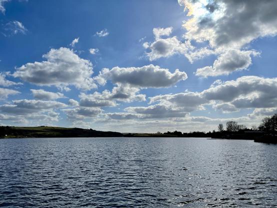 A wide expanse of calm water stretches across the foreground, its rippling surface reflecting the light from a sky dotted with scattered cumulus clouds. The horizon features low rolling hills with patches of green, while the far bank is lined with sparse trees and a few distant buildings silhouetted in the light. The sun, partially obscured by clouds, casts a muted glow and subtle rays across the scene, creating a gentle contrast between the bright sky and the darker water. The atmosphere is tranquil, with no visible human activity on the lake or along the shoreline.