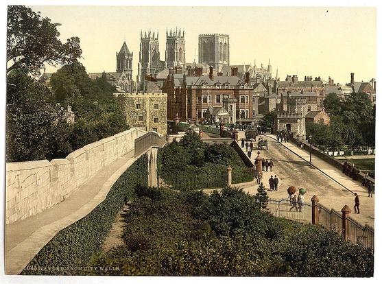 This image depicts a historic urban scene, likely from late 19th to early 20th-century England. It features an elevated vantage point overlooking city walls with a stone parapet and pathways leading into the town below. The lower part of the picture shows well-maintained gardens or greenery along one side of these paths.

A prominent, historical architectural backdrop includes large Gothic-style buildings, which appear to be significant religious institutions such as cathedrals or churches with pointed spires and intricate stonework that characterize this period's ecclesiastical architecture. These structures are set against a clear sky with no visible clouds.

The town below is densely packed with various older stone buildings featuring multi-story facades, dormer windows on the roofs, chimneys, and different architectural details typical of urban British architecture from this era. People can be seen walking along the pathways or standing in groups near streetlights at the lower end of these paths, suggesting a lively public space.

The image has been printed with sepia tones, giving it an aged appearance consistent with early postcards or photographs intended for leisurely appreciation and as keepsakes. The overall ambiance is one of historical charm, urban life from a bygone era, and architectural grandeur reflective of England's cultural heritage during that period.