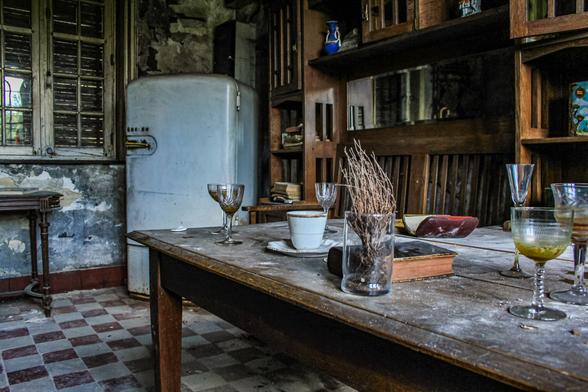Das Bild zeigt ein verlassenes oder vernachlässigtes Interieur mit einem rustikalen Holztisch im Vordergrund. Auf dem Tisch stehen verschiedene Gegenstände wie Gläser, eine Tasse mit Untertasse, ein Glas mit trockenen Zweigen und ein Buch. Im Hintergrund sieht man einen alten Kühlschrank und ein offenes Regal mit Büchern und anderen Gegenständen. Der Raum wirkt heruntergekommen, mit abblätternder Farbe an den Wänden und einem Schachbrettmuster auf dem Boden. Das Tageslicht fällt durch ein vergittertes Fenster und erzeugt eine melancholische Atmosphäre.
