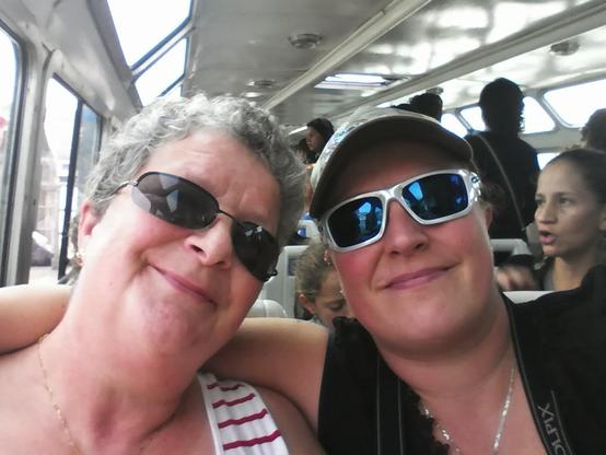 A warm, joyful selfie taken inside a Venice water taxi as it glides from the bus terminal toward Riva degli Schiavoni. On the left sits Cynni’s mum, leaning gently toward her child with a soft, loving smile. She wears dark sunglasses, a white top with red stripes, and a delicate gold necklace, her short curly grey hair catching the daylight filtering through the boat’s large windows. On the right is Cynni, also smiling, wearing reflective white-framed sunglasses, a cap, and a black top, with a camera strap visible around her neck—ready to capture Venice. The two sit close, Cynni’s arm lovingly resting behind her mum, radiating excitement and affection. Around them, other passengers talk and look out at the lagoon, but the focus remains on this happy moment: a mother and her child sharing their anticipation and joy as they head into the heart of Venice for an adventure together.