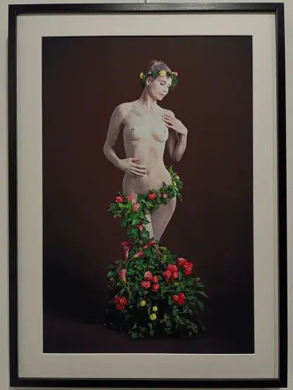 Photographie encadrée montrant une femme nue posant debout, le corps partiellement couvert de feuillage et de fleurs colorées, avec une couronne florale sur la tête, sur fond uni sombre.