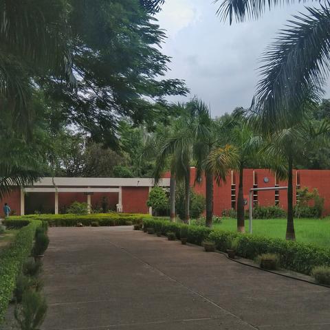 A red brick building is surrounded by lush greenery and tall palm trees under a cloudy sky. A wide walkway leads to the entrance, lined with trimmed bushes.