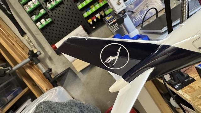 Close-up of a model airplane tail fin with a Lufthansa logo in a workshop setting, surrounded by tools and supplies. 