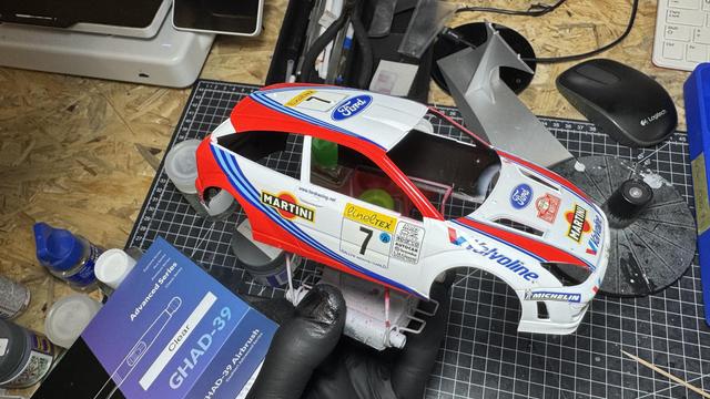 A hand holding a partially assembled and painted model car body with Martini and Ford racing decals on a cluttered workbench.