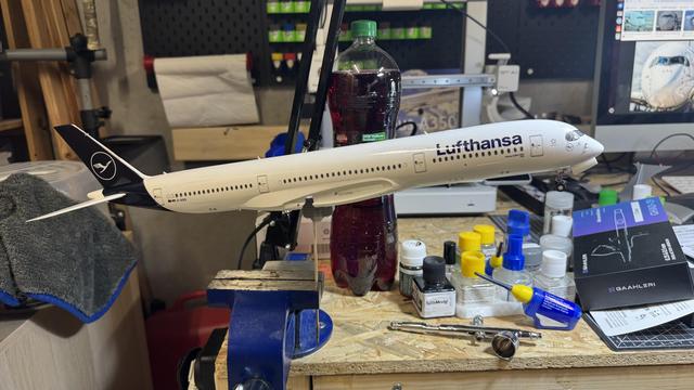A detailed Lufthansa model airplane mounted on a stand on a workbench, surrounded by paint bottles, tools, and a towel in a hobby workspace.