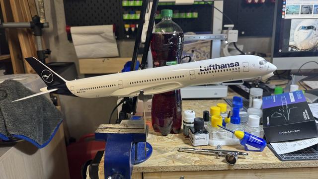 A detailed Lufthansa model airplane mounted on a stand on a workbench, surrounded by paint bottles, tools, and a towel in a hobby workspace.