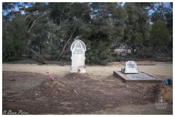 A photograph of a solitary grave in the Kingston Cemetery. In the foreground, a mound of reddish brown dirt marks a grave, topped with a single pink flower.  Directly behind this, a narrow, weathered stone headstone with a distinct pointed arch (Gothic) top stands upright, bearing faded inscriptions.  To the right, a smaller, rectangular granite monument is set within a bordered plot covered in light gravel. The cemetery ground is primarily dry, dark soil.  The background is dense with dark green to brown native scrub and trees, creating a shaded and isolated setting.