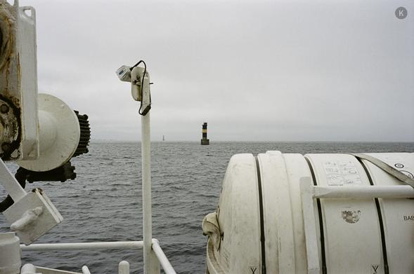 Blick vom Deck eines Schiffs über eine graue, leicht wellige See; im Vordergrund weiße Rettungsbojen und Metallausrüstung, in der Ferne ein schlanker Leuchtturm unter bedecktem Himmel.