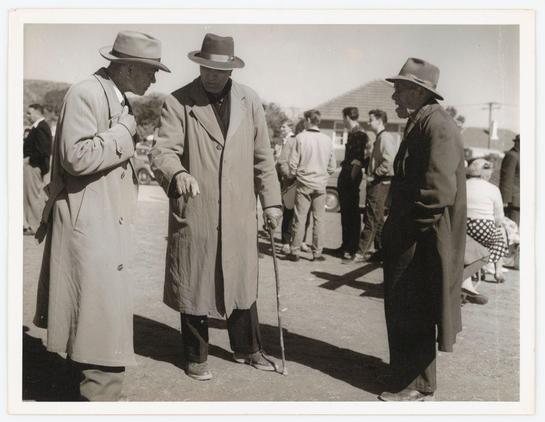 The photograph captures a moment from what appears to be an outdoor gathering or event, likely during the mid-20th century given its black and white composition. Three men are prominently featured in the foreground; all of them wear hats—a fedora for two on one side and a wide-brimmed hat for another—and long coats suitable for cool weather. The man with the cane is engaged in conversation with his left hand gesturing towards something outside the frame, while standing next to him looks down at what could be an object or document he's holding. Their relaxed postures suggest familiarity among themselves.

In the background, there are several more individuals mingling around—a mix of men and women wearing period-appropriate attire—indicating a social setting like a fair, community event, perhaps even sports-related given one man’s posture that could be interpreted as walking or carrying something not entirely visible in this frame. The environment is bright with natural sunlight casting shadows on the ground, hinting at daytime hours.

This scene provides insight into historical fashion and societal gatherings during its time period. It doesn't offer clear evidence about a specific location but seems like an everyday public event of considerable interest to those who attended.