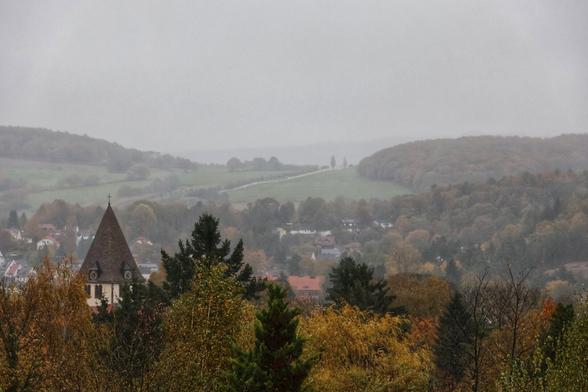 A misty, autumnal landscape showcasing a quaint village nestled among rolling hills and dense woodland. In the foreground, the pointed steeple of a church rises above the treetops, its dark roof contrasting with the muted colours of the season. The village below is dotted with houses and buildings, partially obscured by a light fog that softens the scene. The hills in the background are covered in patches of green fields and clusters of trees, their foliage displaying hints of autumnal browns and oranges. The overall atmosphere is calm and serene, evoking a sense of peaceful rural life.