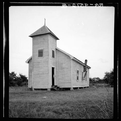 The photo captures a solitary wooden chapel situated in an open field. The structure is simple, featuring plain white walls and basic construction with no ornamental details, suggesting it serves the community as more of a functional space than one for elaborate services or ceremonies.

It's evident that this church has seen better days; weathering on its siding indicates age and possible neglect. A prominent feature is an old wooden ladder attached to the side, leaning against what seems like a support beam, which suggests either maintenance work in progress or perhaps decayed wood requiring replacement.

The chapel lacks windows on most of its sides, with only one visible window beside the entrance and another above near the roofline. The pitched gable ends are topped with simple crosses, adding to the simplicity yet distinctiveness of this rural place of worship.

Surrounding vegetation is sparse but includes some bushes or small trees that appear wild and unmanaged, further hinting at a setting where human intervention isn't frequent. No people are visible in the shot, emphasizing solitude. The backdrop is an overcast sky with no discernible landscape features other than distant foliage, suggesting isolation.

The monochrome tone of this black-and-white image adds to its historic ambiance, evoking nostalgia and possibly hinting at a bygone era.